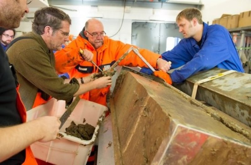 Polarstern expedition leader Prof Dr Rüdiger Stein (center, in orange clothing) and his team examine the salvaged sediment core from the Lomonosov Ridge aboard the Polarstern lab. (Photo: Alfred Wegener Institute / Audun Tholfsen, UoB)